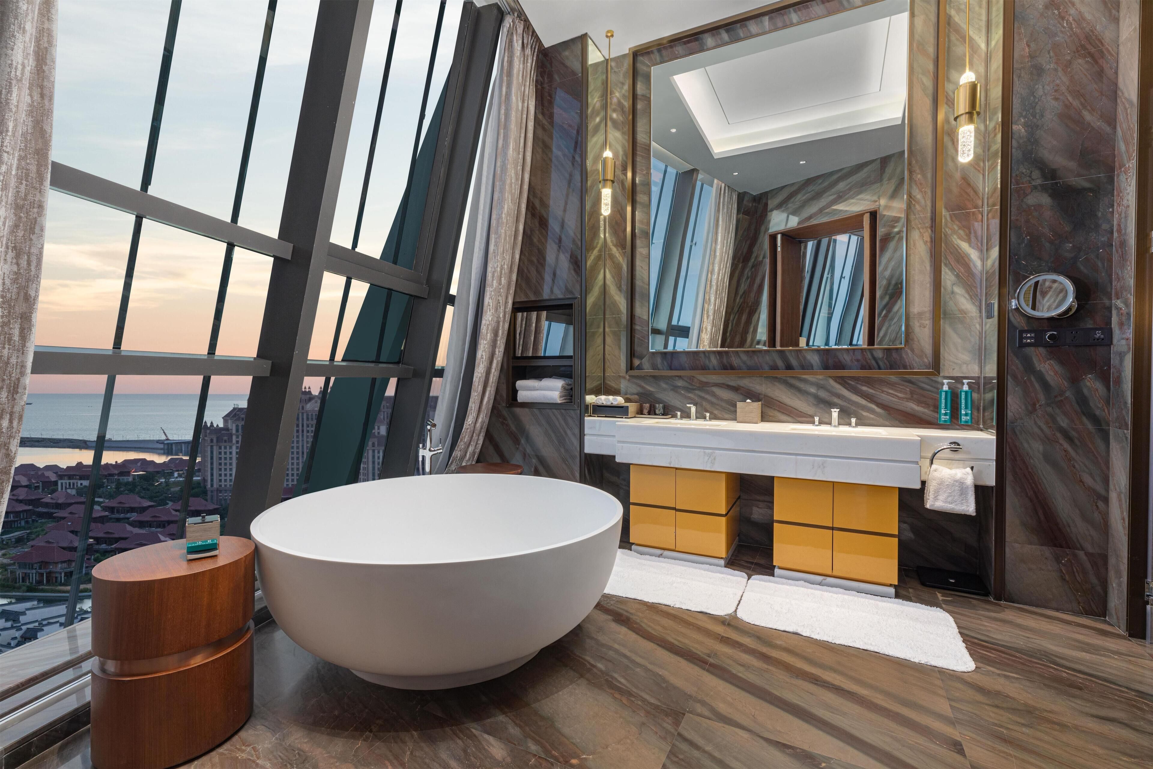 Un baño moderno con una bañera grande, un tocador con espejo y vistas de la ciudad a través de amplias ventanas.