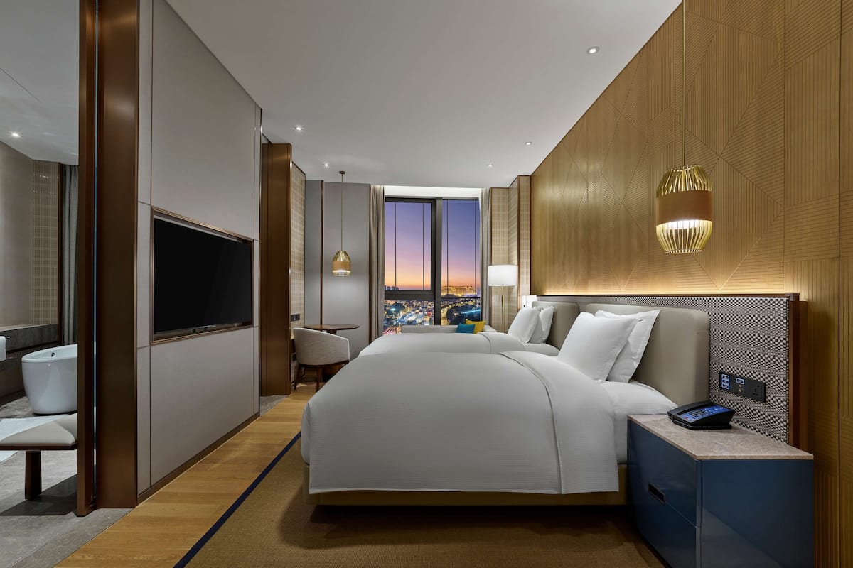 Habitación de hotel moderna con una cama grande, televisión, escritorio y vistas a la ciudad al atardecer.