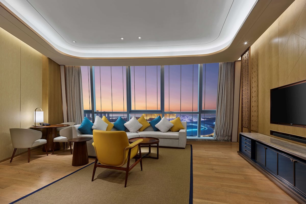 Una habitación de hotel moderna con un ventanal grande, un sofá con cojines de colores, un sillón amarillo, una mesita y un televisor de pantalla plana.