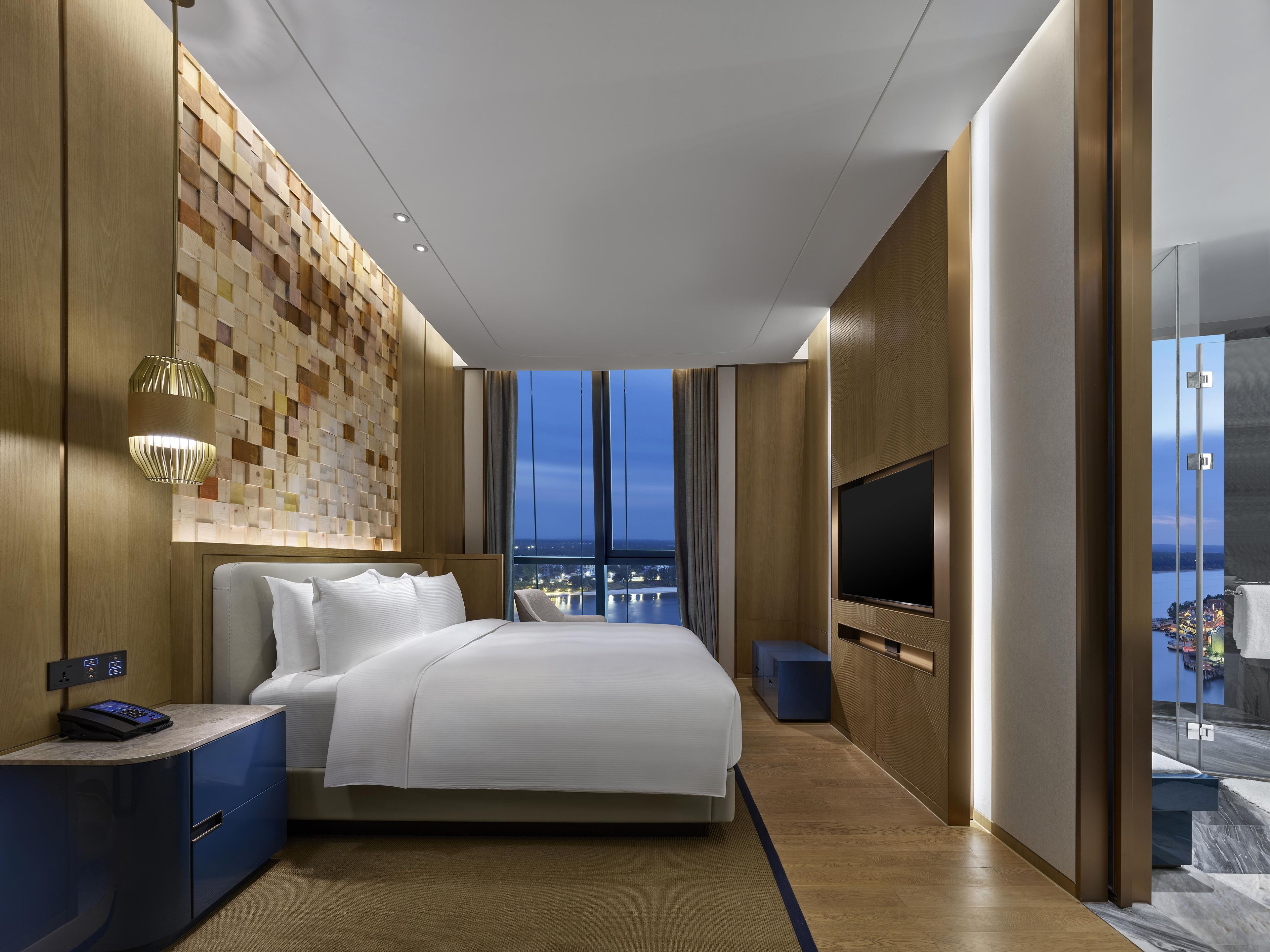 Una habitación de hotel moderna con una cama grande, un televisor, un escritorio con panel de control y vistas a la ciudad a través de las ventanas.