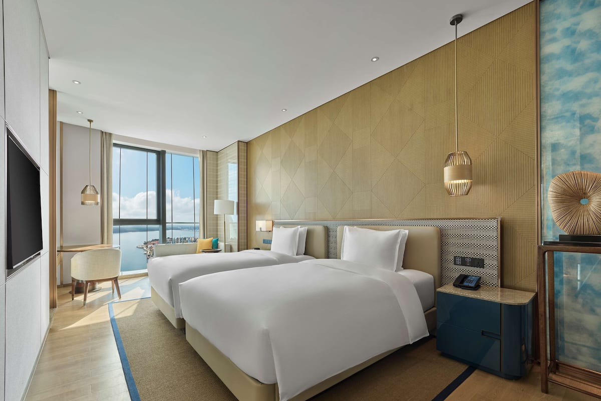 Una habitación de hotel moderna con dos camas grandes, un escritorio, una silla, un ventanal amplio con vista a la ciudad y un panel decorativo en la pared.