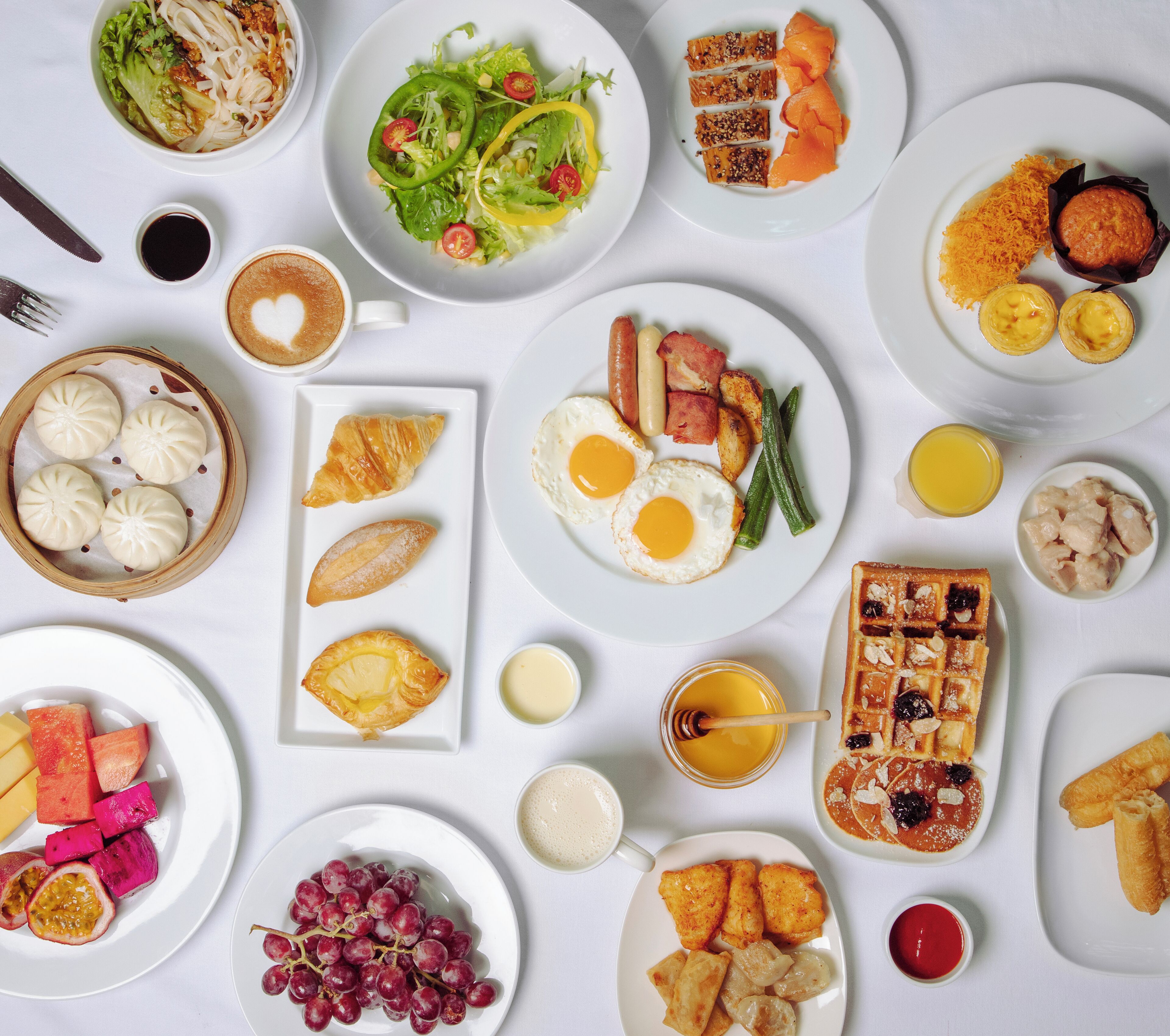 Variedad de platos para el desayuno, como huevos, salchichas y bollería, sobre una mesa blanca.