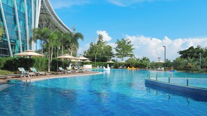 A swimming pool with lounge chairs and umbrellas, surrounded by palm trees and a modern building. - Hilton Hainan Ocean Flower Island (Danzhou)