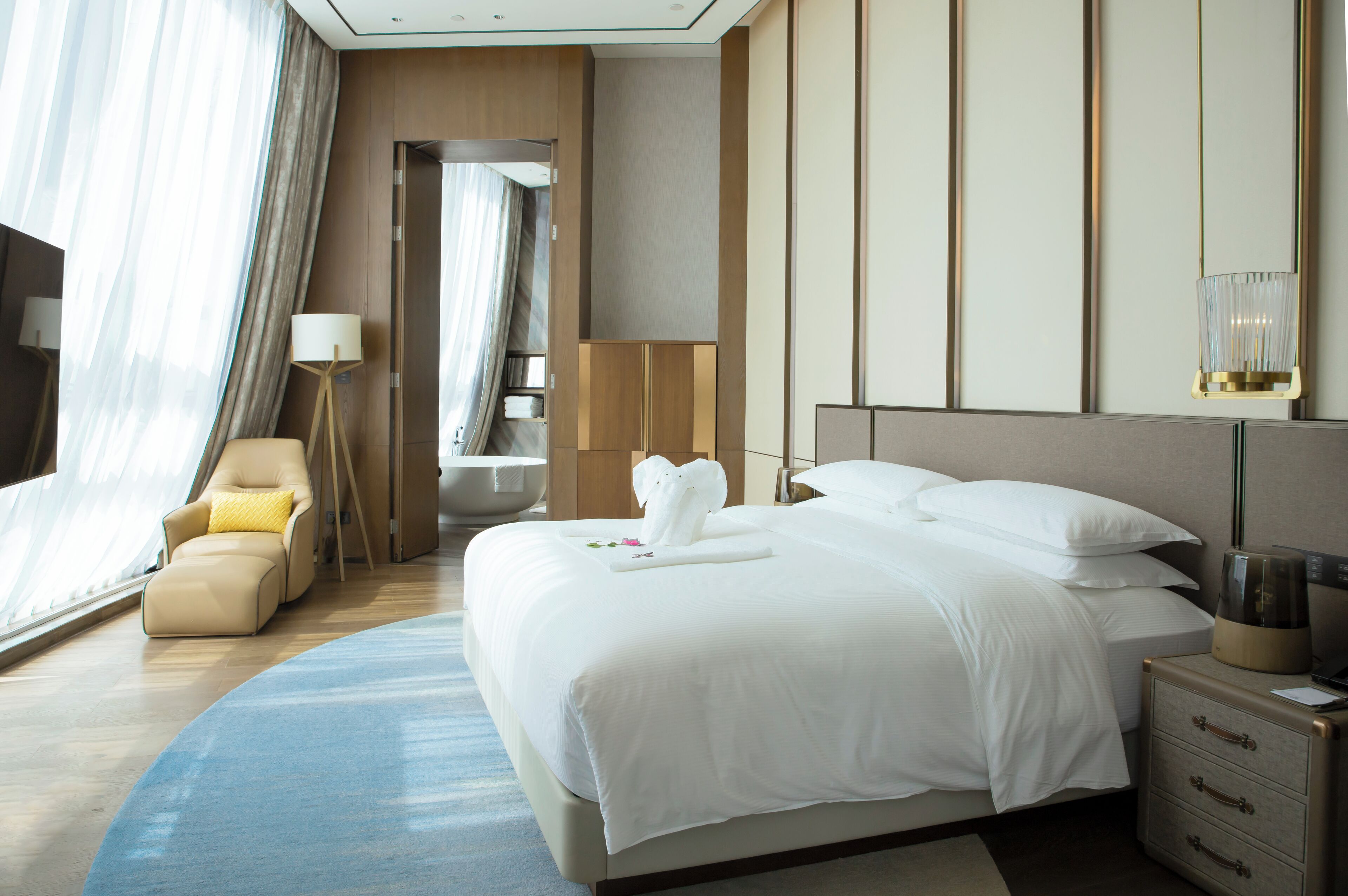 A modern hotel room with a large bed, a sitting area with a chair, a bedside table with a lamp, and a view of the outside through large windows.