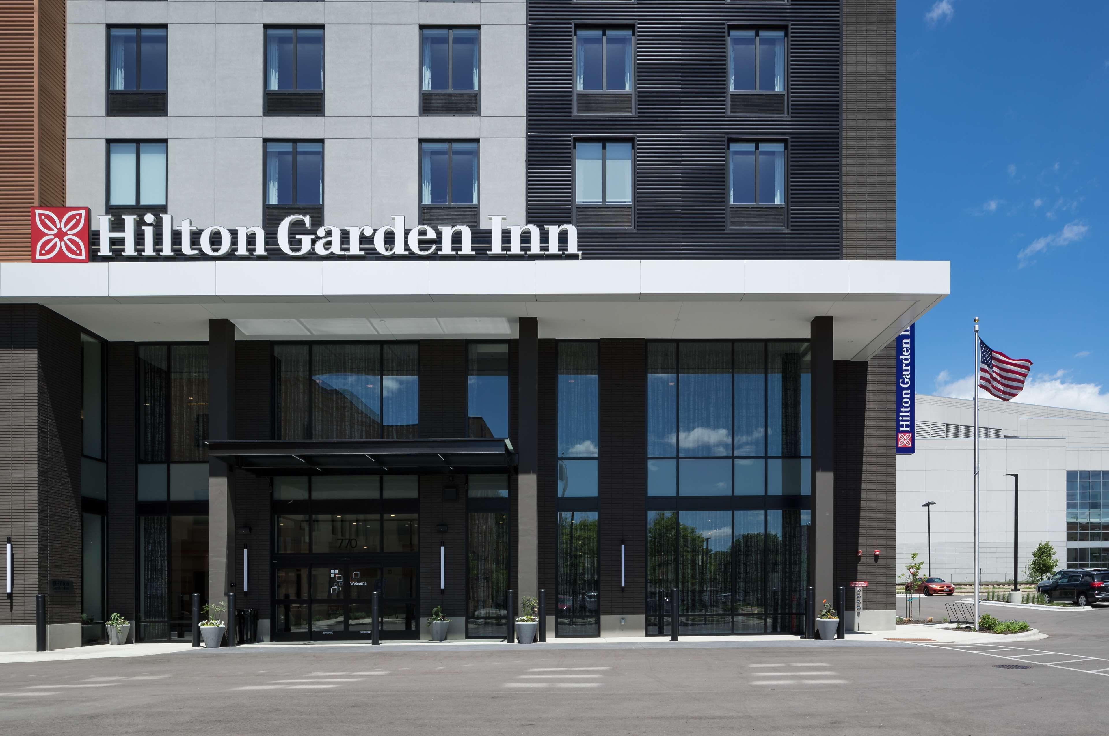 Exterior de un hotel Hilton Garden Inn con diseño moderno, ventanas grandes y una bandera estadounidense.