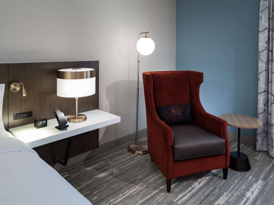 A hotel room with a bed, a desk with a lamp, a red armchair, a small table, and a patterned curtain.