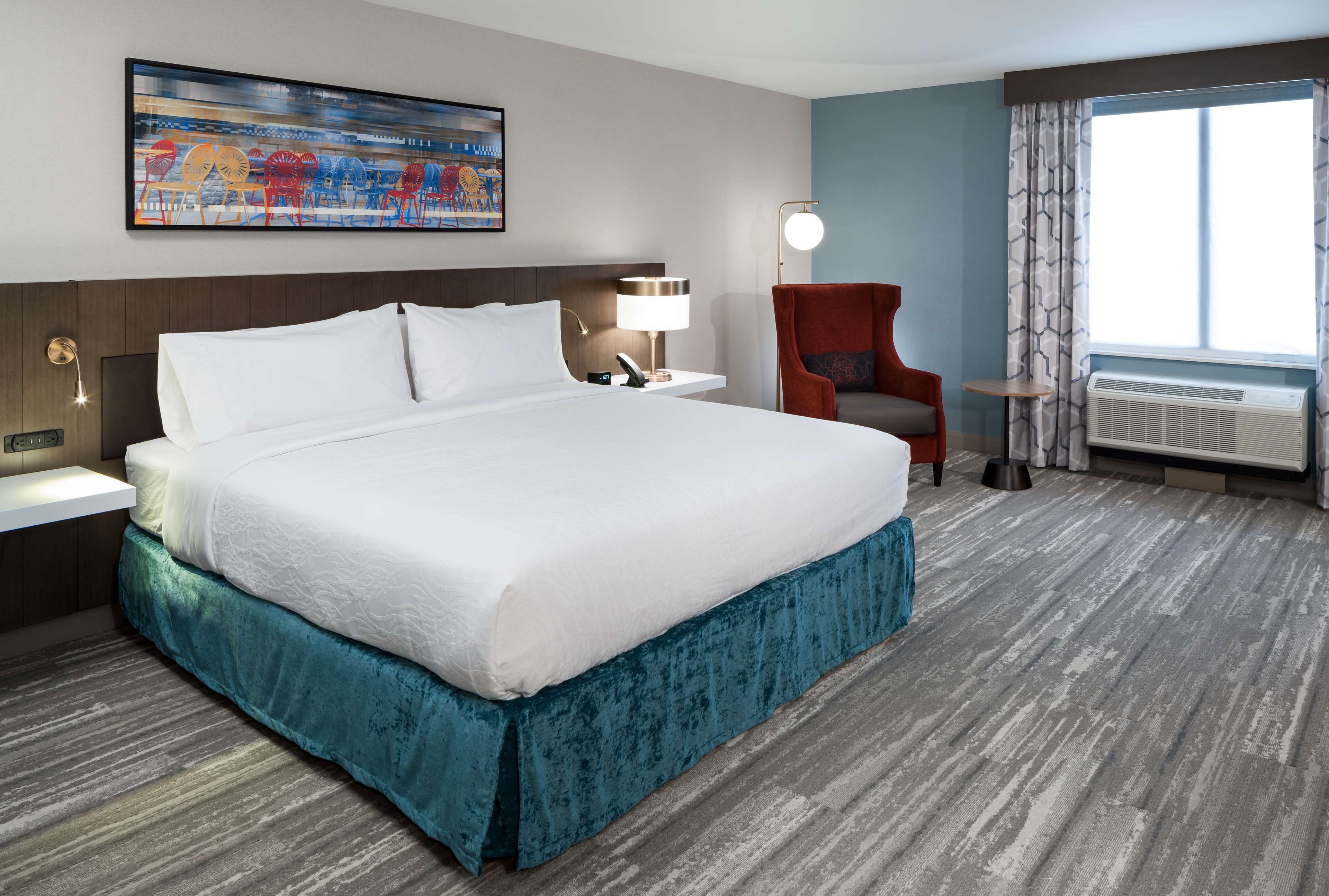 Quarto de hotel com uma cama grande, mesa de cabeceira com abajur, uma cadeira, uma pequena mesa e uma janela com cortinas.