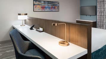 A hotel room with a desk, chair, bed, TV, and artwork on the wall.