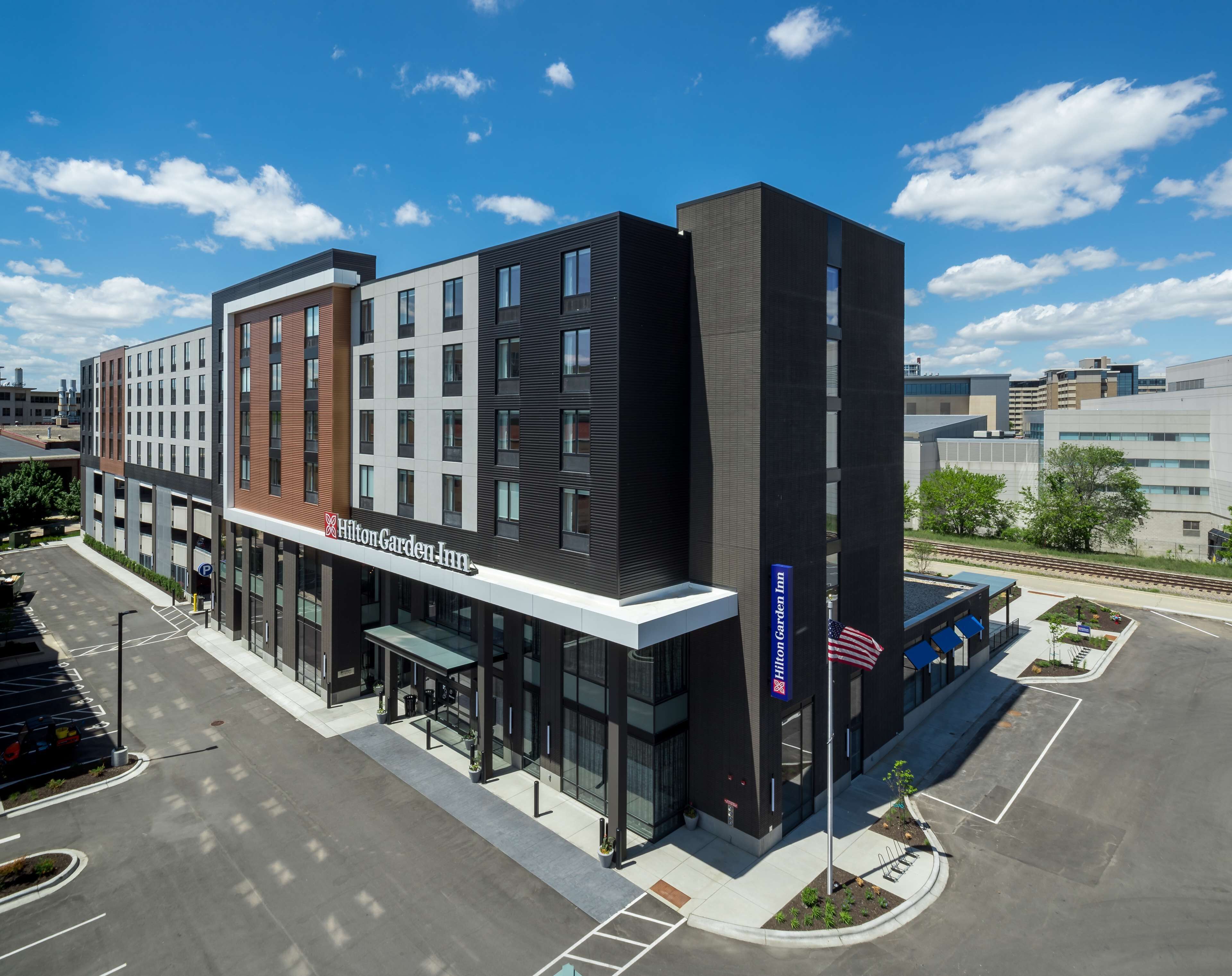 A modern hotel with a prominent Hilton Garden Inn sign, a parking lot, and a flagpole with an American flag.