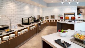 A modern kitchen with white tiled walls, stainless steel appliances, and a breakfast bar with fruit and beverages. - Home2 Suites by Hilton Riverside March Air Force Base (March Air Reserve Base)