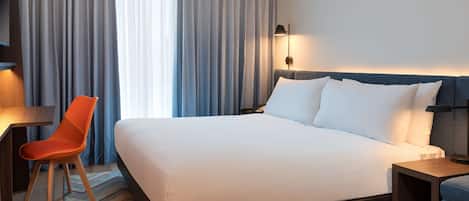 A hotel room with a large bed, a desk, an orange chair, and a nightstand with a lamp.