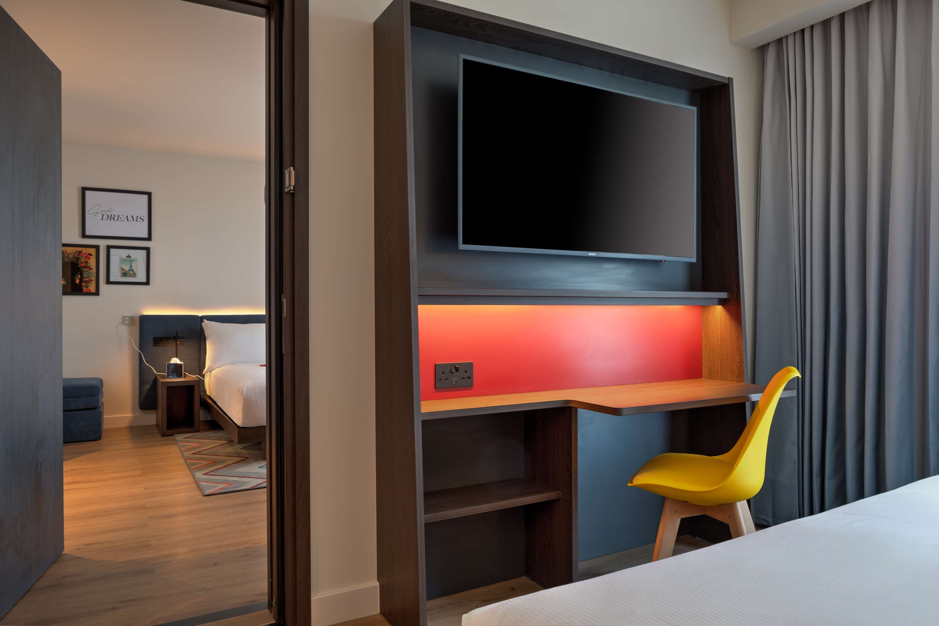 Ein Hotelzimmer mit Bett, Schreibtisch mit Stuhl, einem an der Wand befestigten Fernseher und einem kleinen Nachttisch mit Lampe.