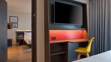 A hotel room with a bed, a desk with a chair, a TV mounted on the wall, and a small bedside table with a lamp.