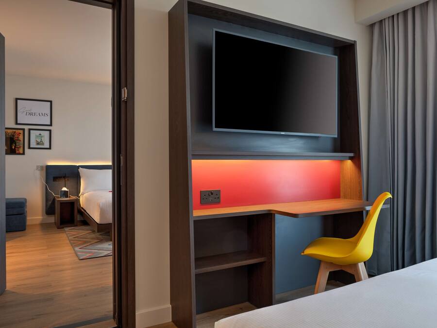 A hotel room with a bed, a desk with a chair, a TV mounted on the wall, and a small bedside table with a lamp.