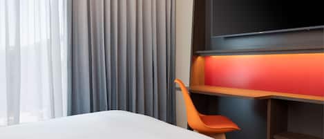 A hotel room with a bed, a TV mounted on the wall, a desk with a red light, and a chair.