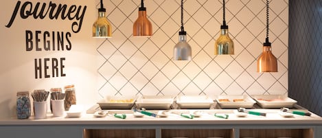 A buffet setup with various food items and utensils, a wall with a motivational quote, and hanging pendant lights.