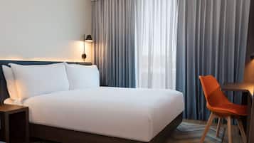 A hotel room with a large bed, a desk with a chair, a lamp, and a view through sheer curtains.