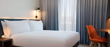 A hotel room with a large bed, a desk with a chair, a lamp, and a view through sheer curtains.