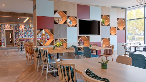 Een moderne eetruimte met houten tafels en stoelen, een grote flatscreen televisie en kleurrijke geometrische wanddecoraties.