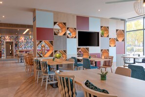 A modern dining area with wooden tables and chairs, a large flat-screen TV, and colorful geometric wall decorations.
