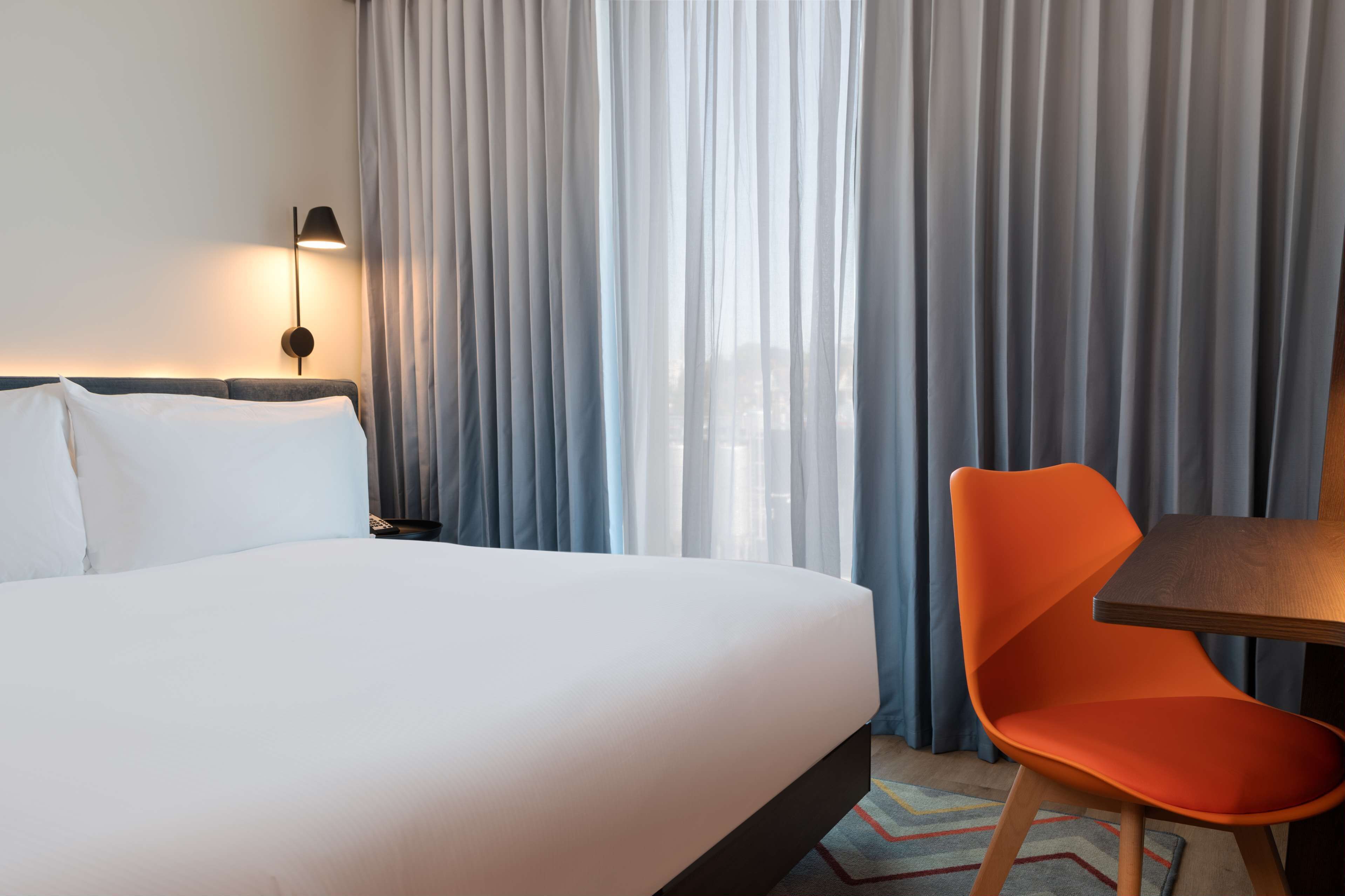 A hotel room with a bed, a chair, a table, and a lamp.