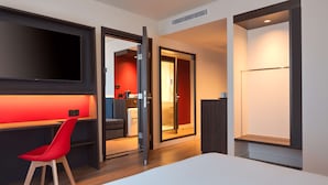 Een moderne hotelkamer met een groot bed, een bureau met een rode stoel, een aan de muur bevestigde televisie en een schuifdeur naar een badkamer.