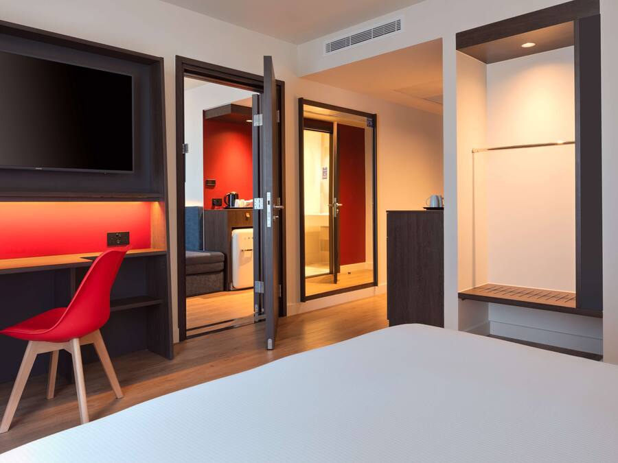 Une chambre d’hôtel moderne avec un grand lit, un bureau avec une chaise rouge, un téléviseur fixé au mur et une porte coulissante donnant accès à la salle de bain.