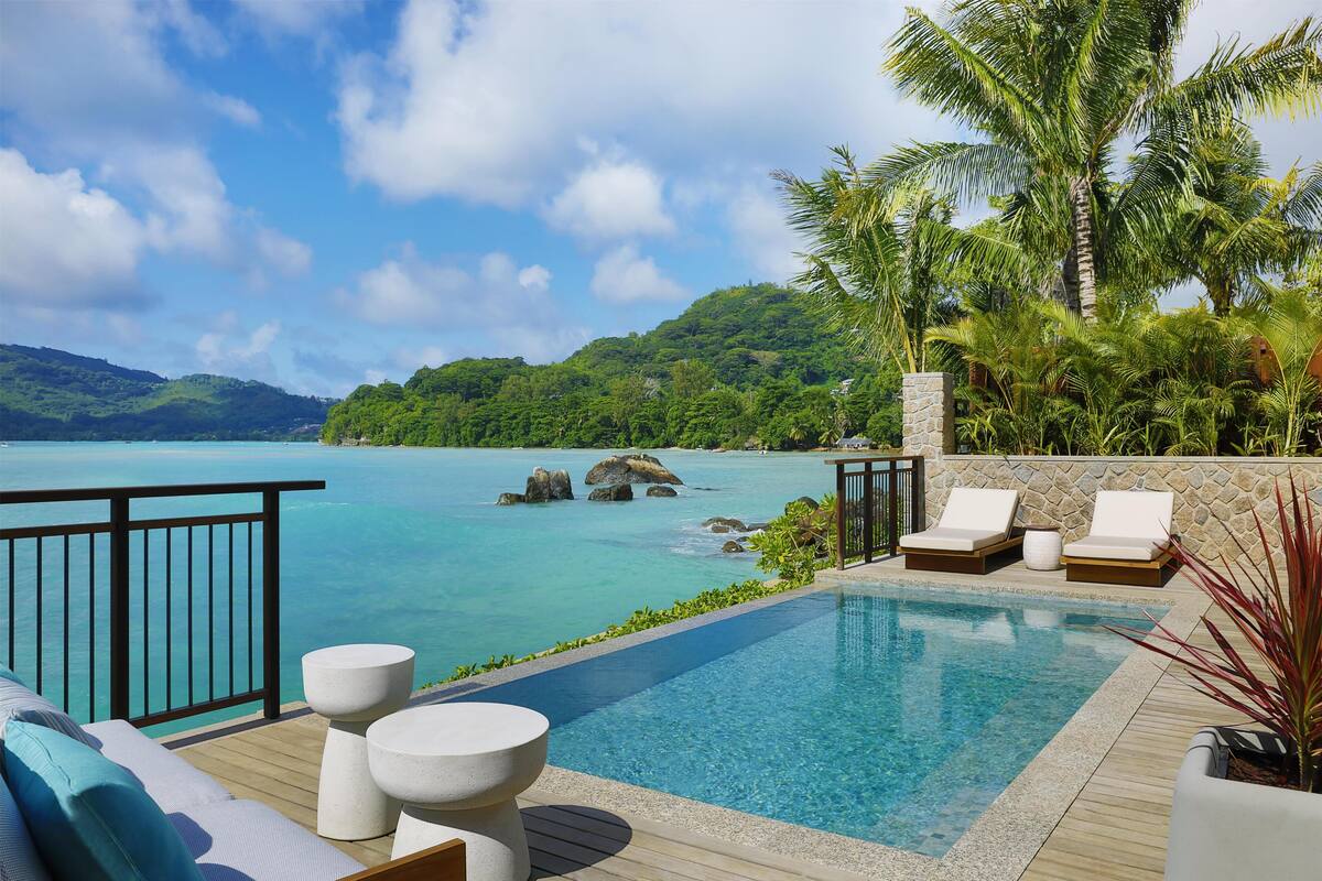 A pool with a view of the sea and mountains, lounge chairs, and a wooden deck.