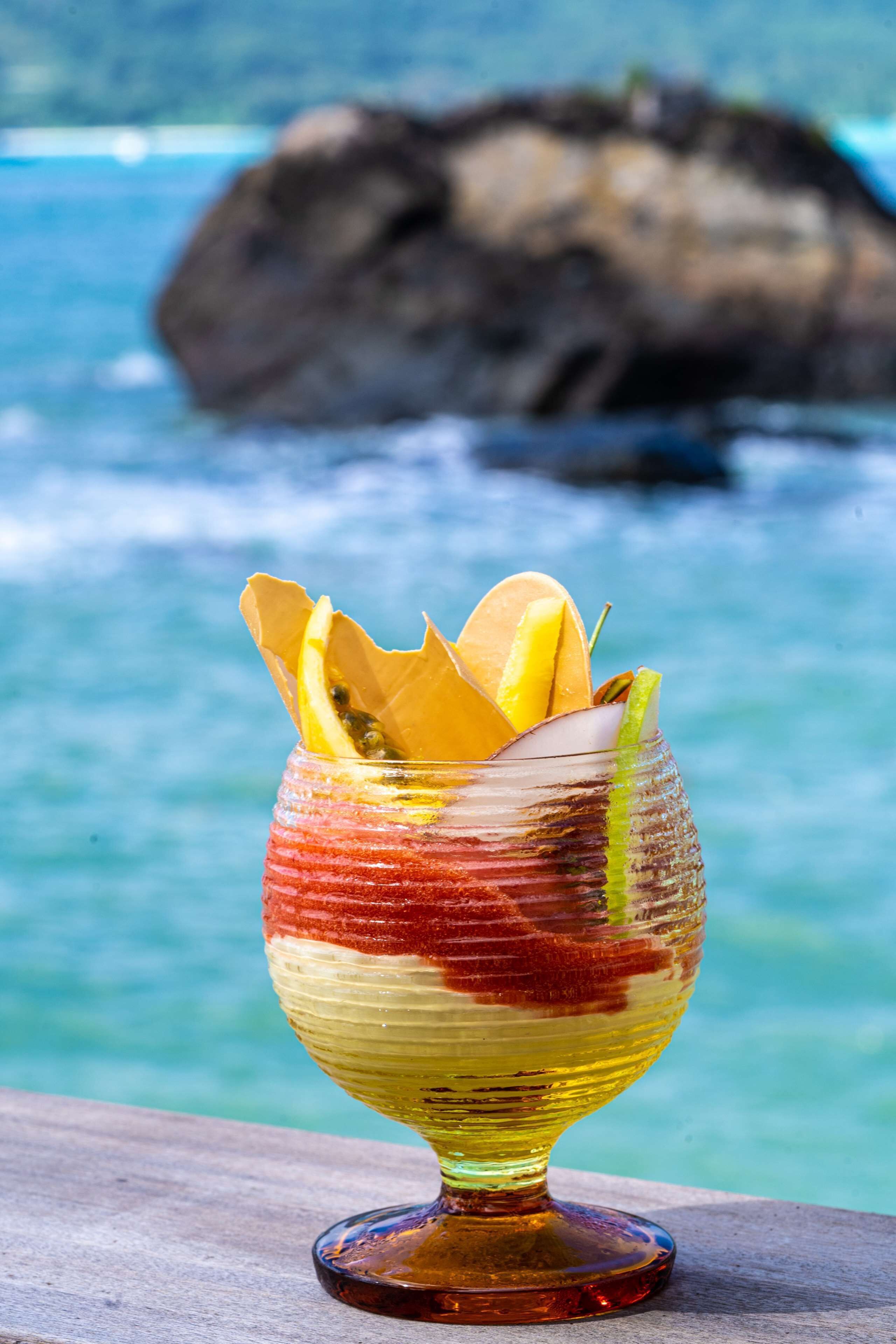 Una copa de cóctel con guarnición de frutas sobre una superficie de madera, con una costa rocosa y agua turquesa al fondo.