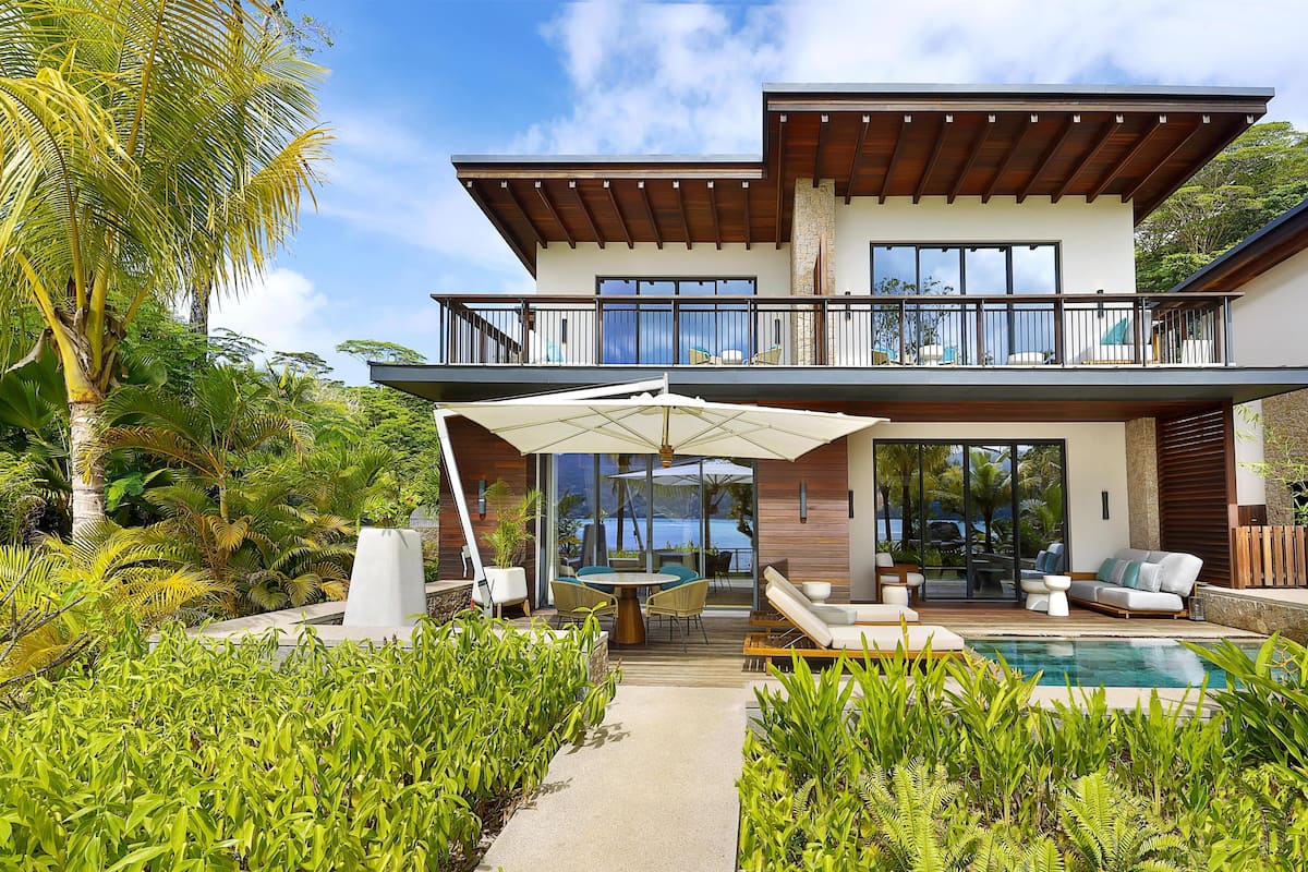 A two-story modern house with a balcony, a pool, and lush tropical vegetation.