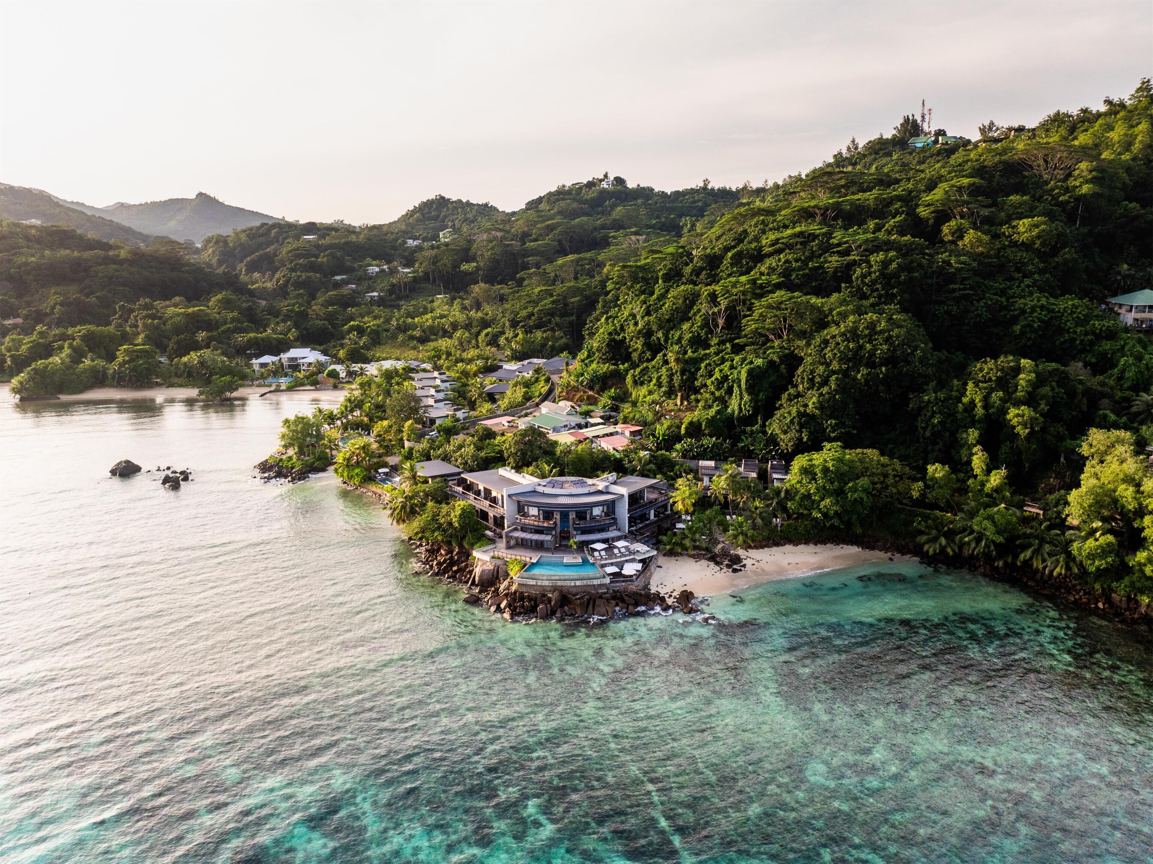 An aerial view of a coastal resort with villas, a swimming pool, and lush greenery.