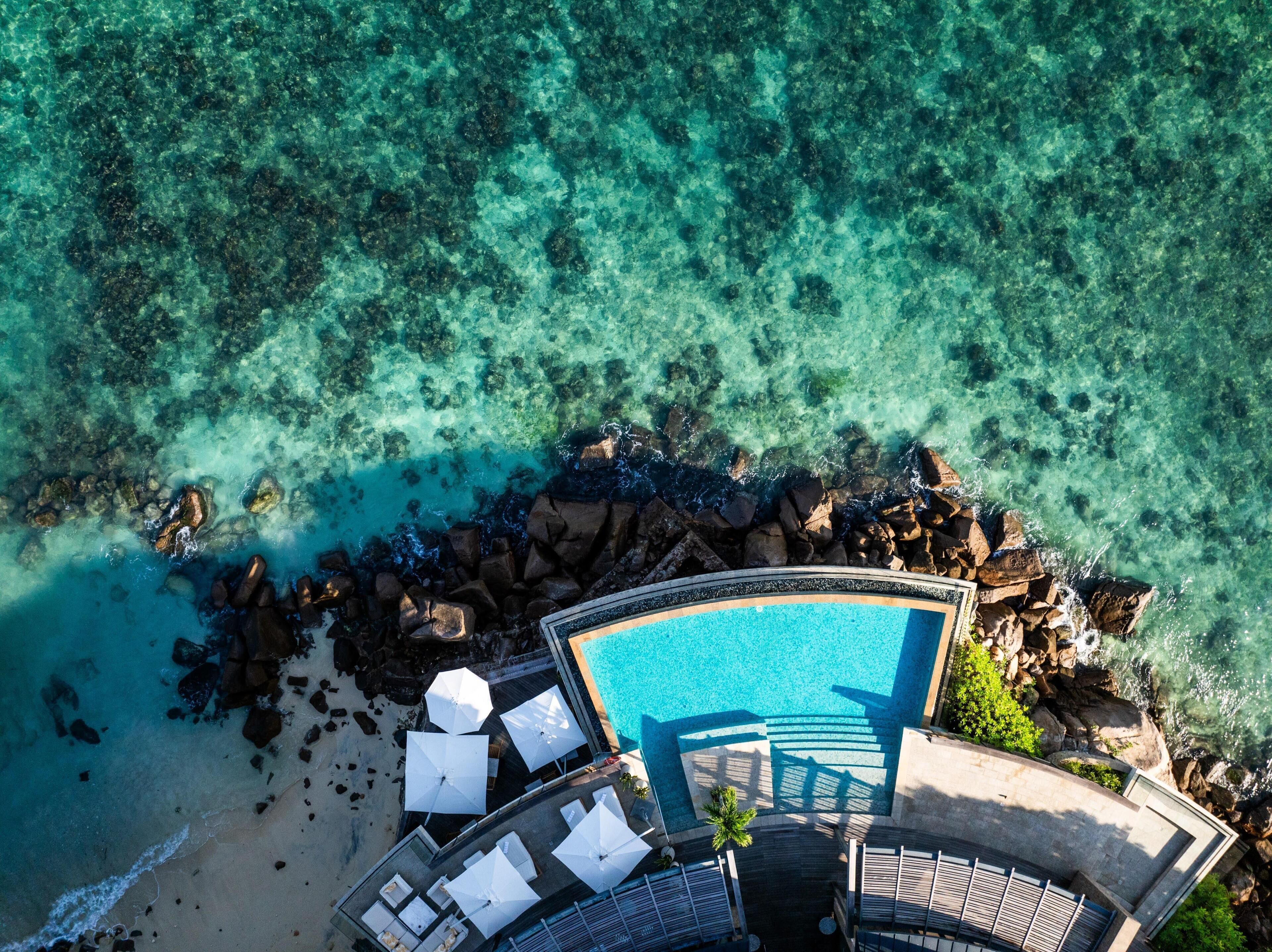 An aerial view of a resort with a clear pool, lounge chairs, and a rocky shoreline.