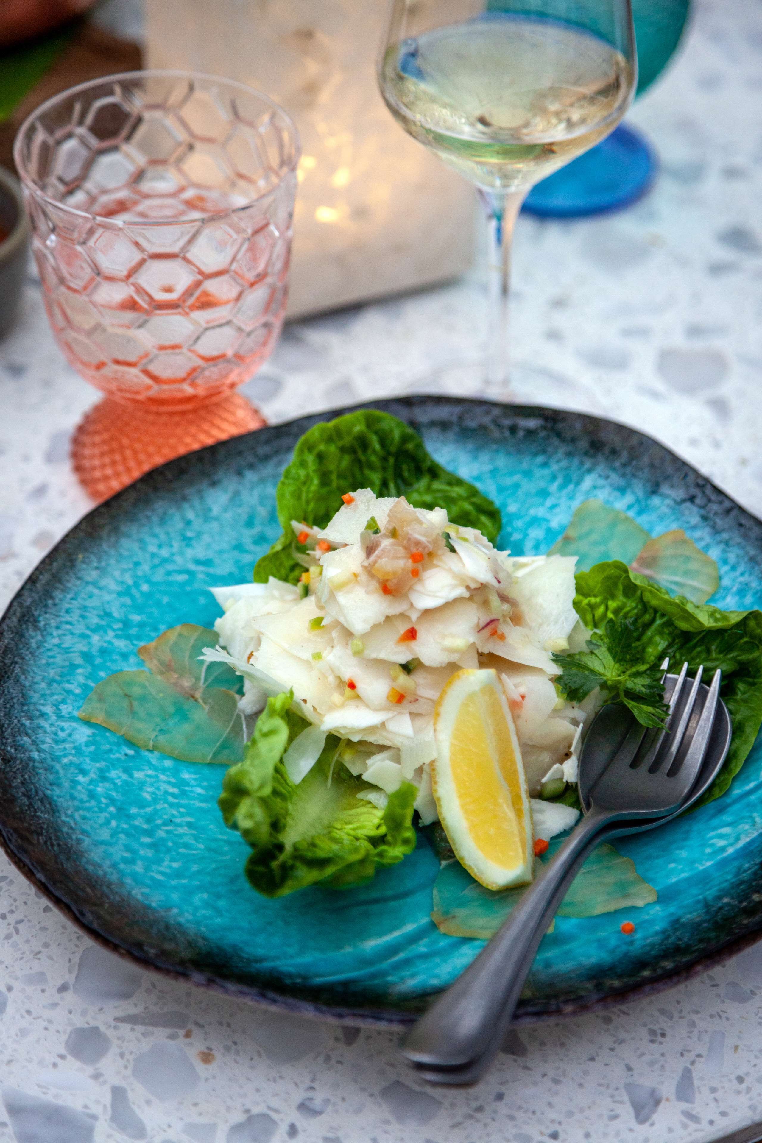 A plate of ceviche with lemon and lettuce on a blue plate, accompanied by a glass of white wine and a pink glass.
