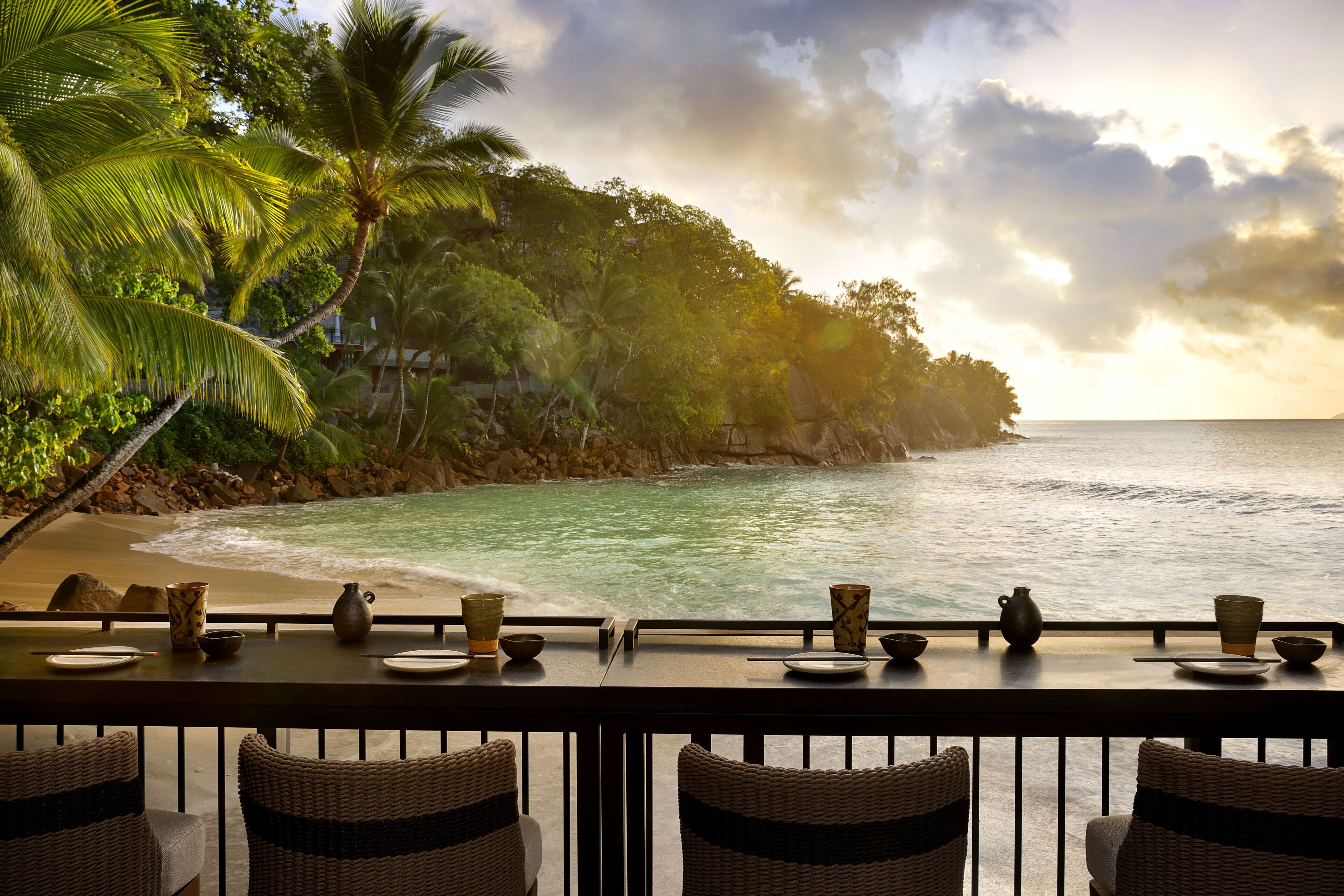 Un área de comedor junto al mar con sillas de mimbre y una mesa dispuesta con platos, tazones y tazas, con vista a una playa con palmeras y el océano.