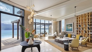 A spacious living area with a large window overlooking a beach, a modern fireplace, and a bookshelf filled with books. - Mango House Seychelles, LXR Hotels & Resorts (Mahé Island)