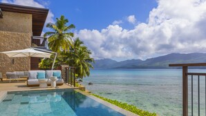 Ein Poolbereich mit Blick auf das Meer und die Berge, eine Lounge mit Sofa und Sonnenschirm sowie ein Steinhaus.