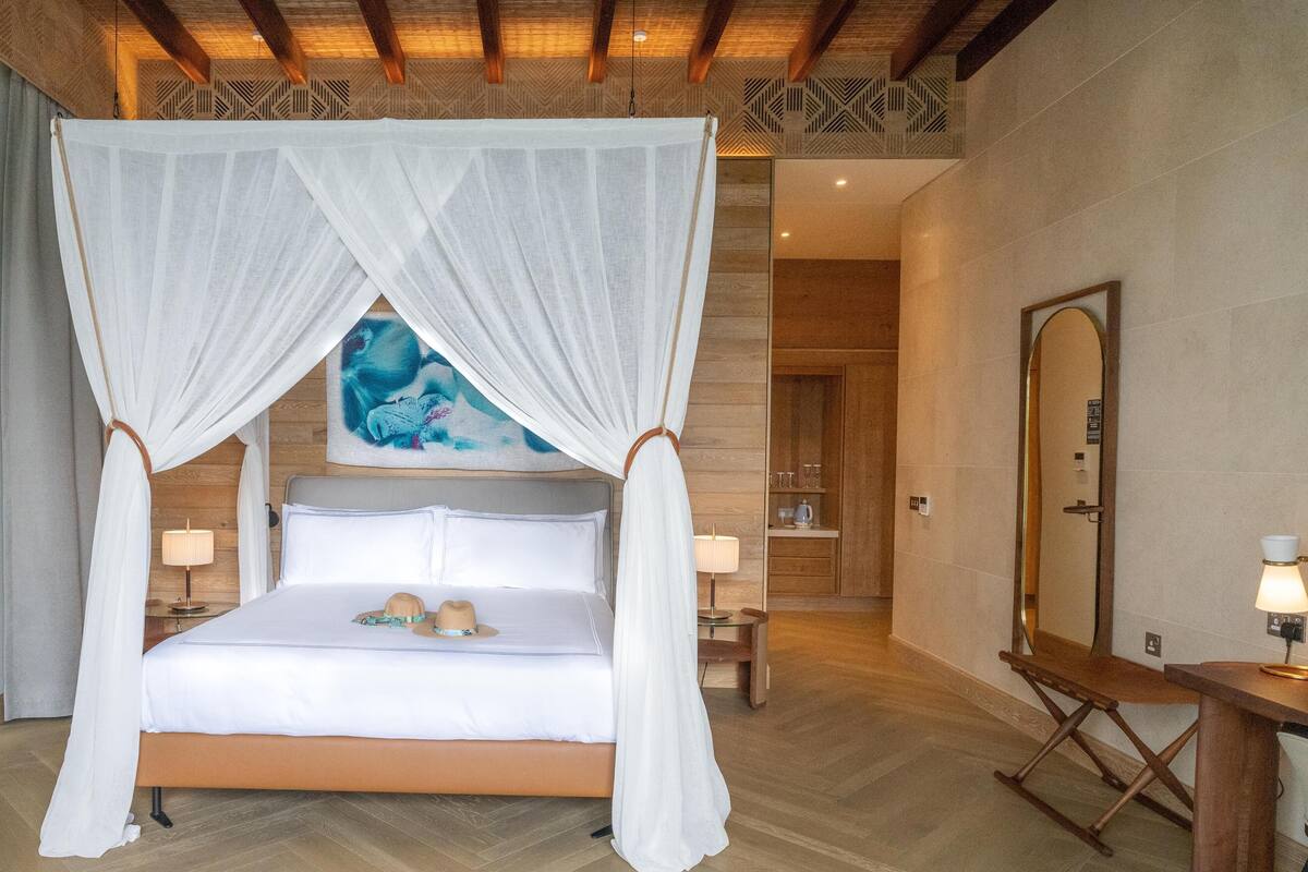 A bedroom with a four-poster bed, a bedside table with a lamp, a wooden bench, and a large mirror.
