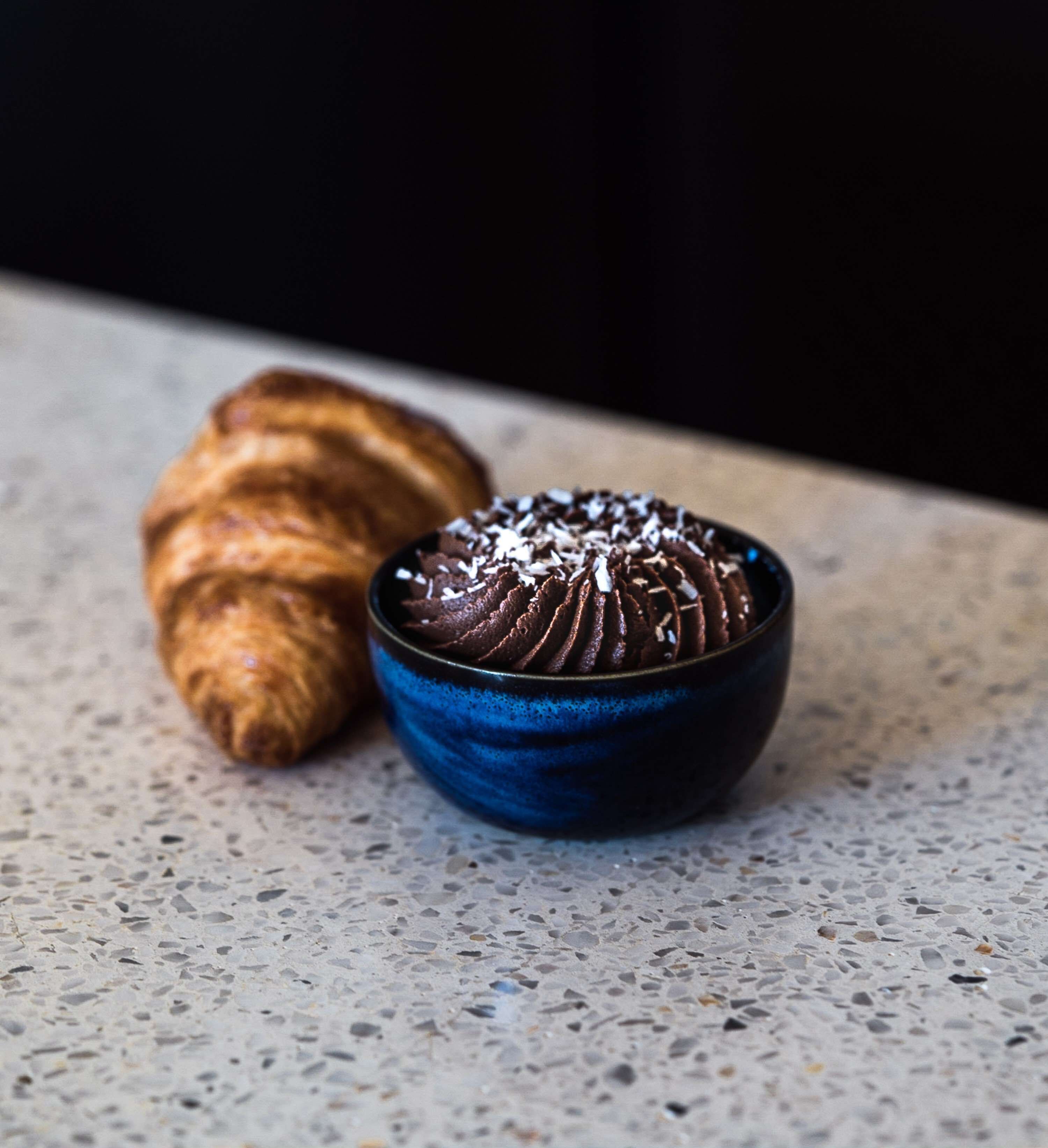 A croissant and a bowl of chocolate mousse on a speckled countertop.