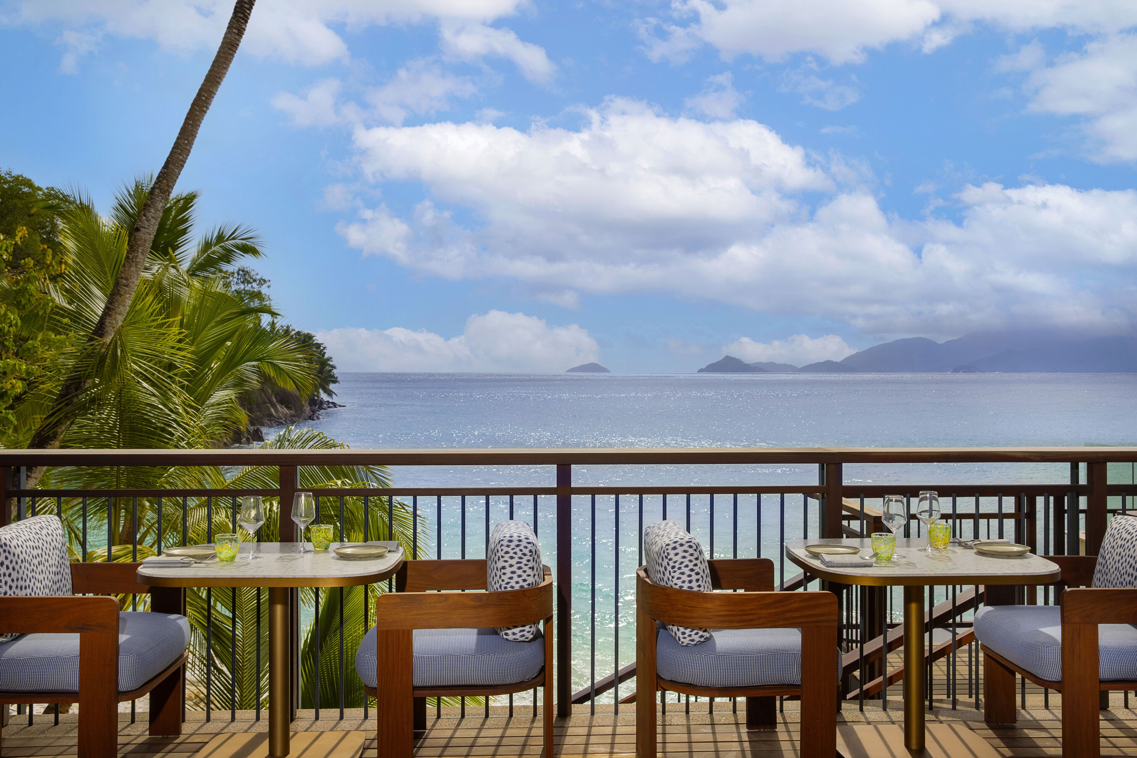 Terraço com duas mesas, cada uma com quatro cadeiras, com vista para uma praia com palmeiras e o oceano.