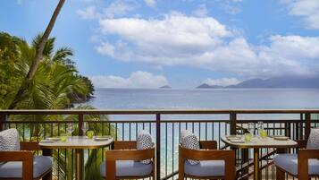 Terraço com duas mesas, cada uma com quatro cadeiras, com vista para uma praia com palmeiras e o oceano.