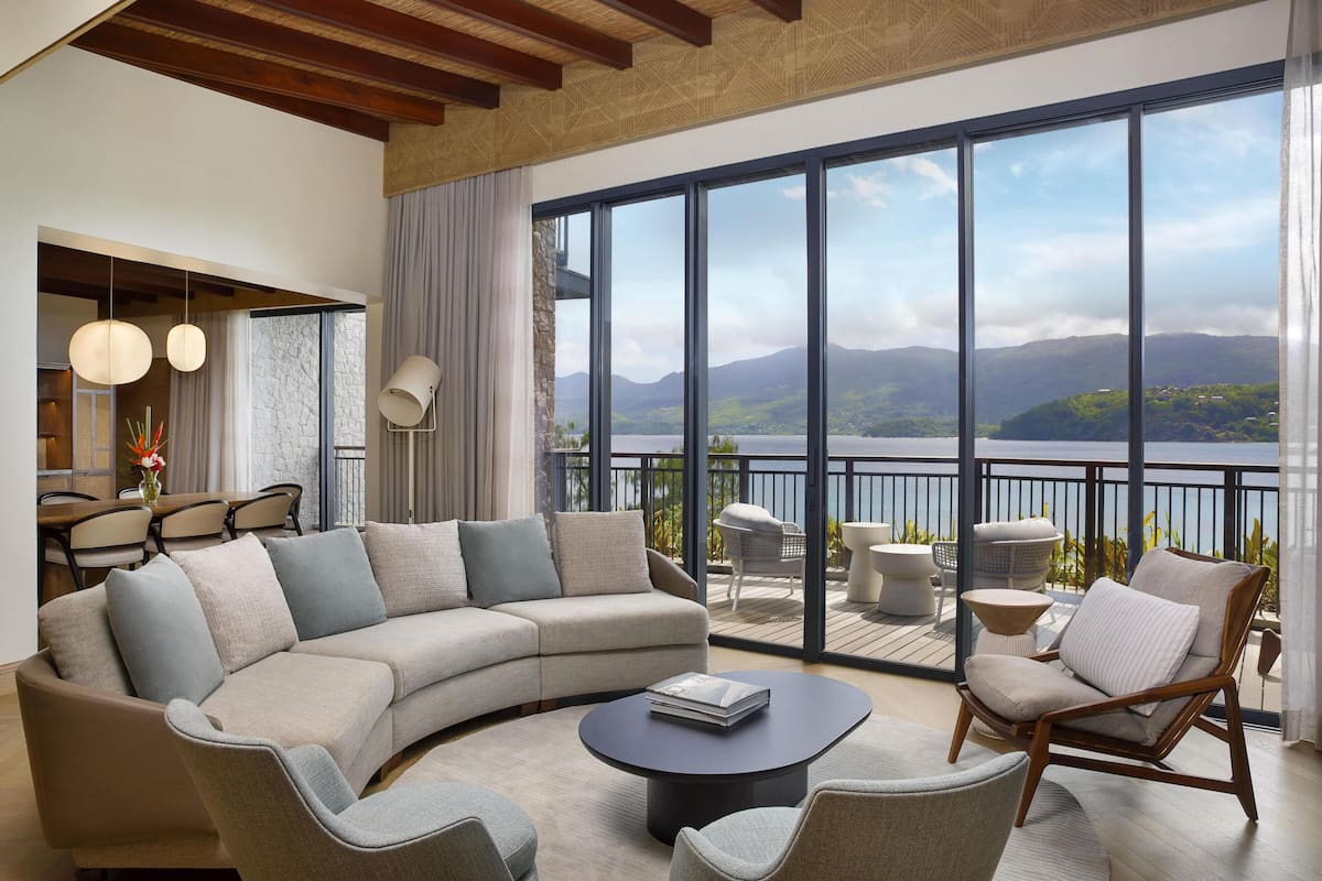 A modern living room with a curved sofa, a coffee table, and a view of a lake and mountains through large windows.