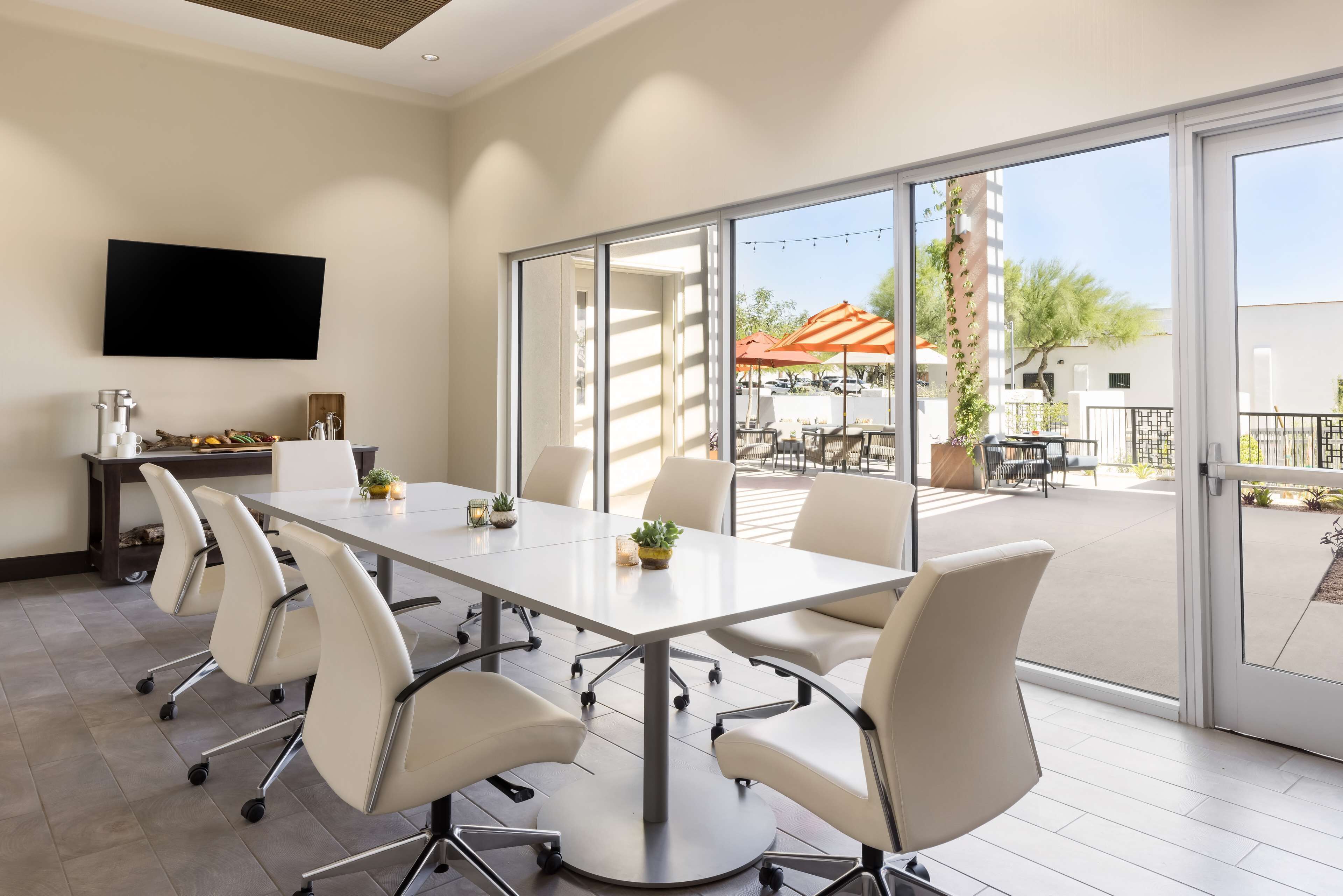 A modern meeting room with a large table, white chairs, a flat-screen TV, and a view of an outdoor patio area with umbrellas.