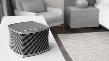A modern, minimalist room with a grey, cylindrical air purifier on a white table, a grey sofa with a patterned cushion, a white bedside table with a lamp, and a grey bed with white bedding.