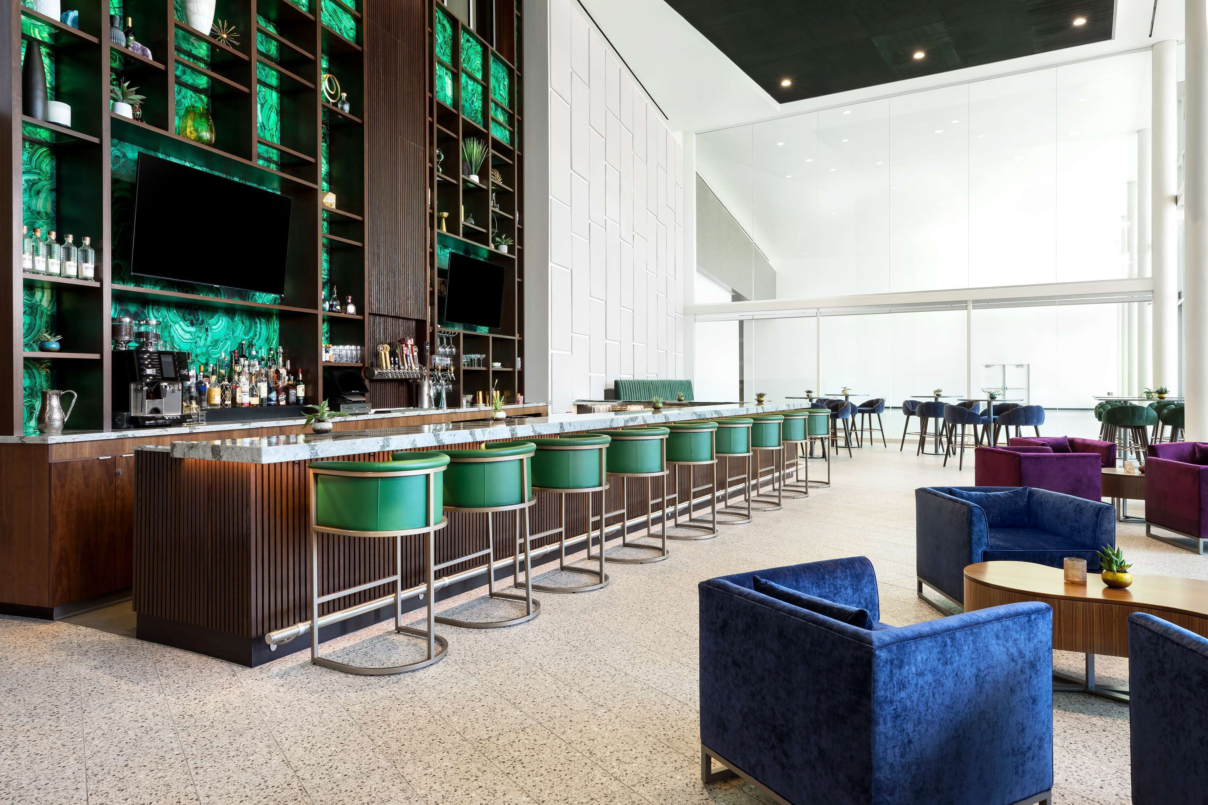A modern bar area with a long counter, green bar stools, and a variety of bottles and glasses. There are several seating areas with sofas and tables, and a large window providing natural light.