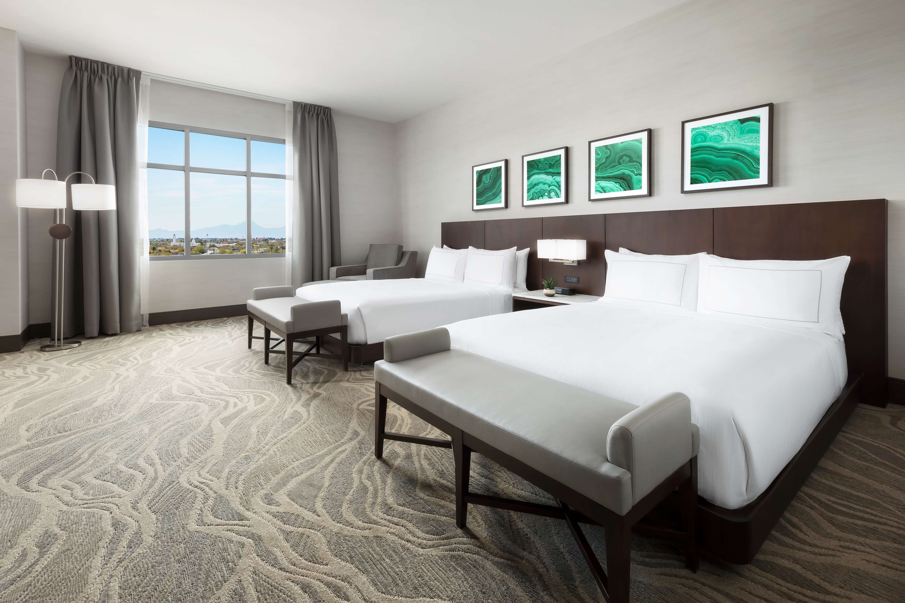 A hotel room with two beds, a desk, a chair, a large window with a view, and three framed artworks on the wall.