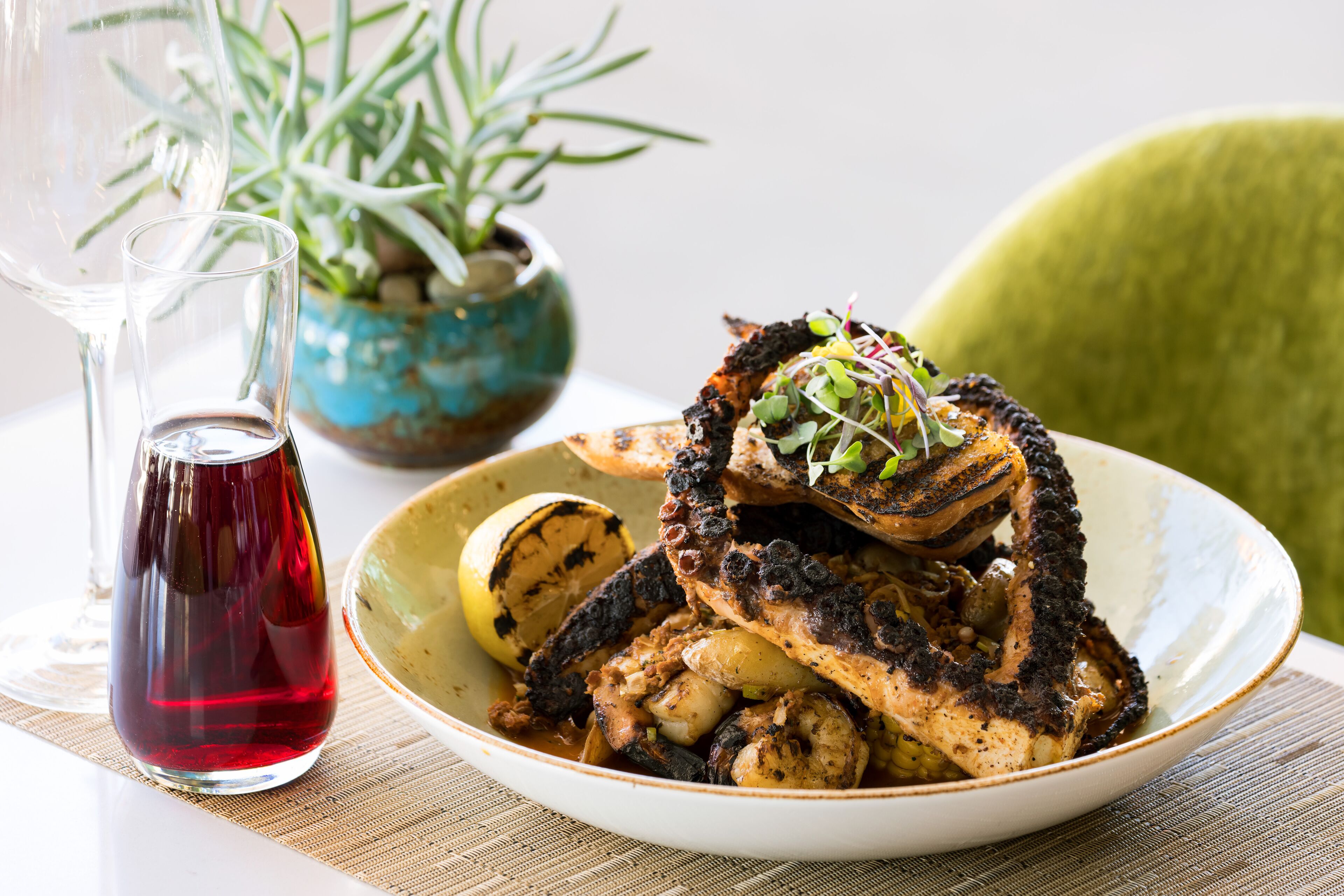 A plate of grilled chicken with vegetables and a glass of red wine on a table with a plant in the background.