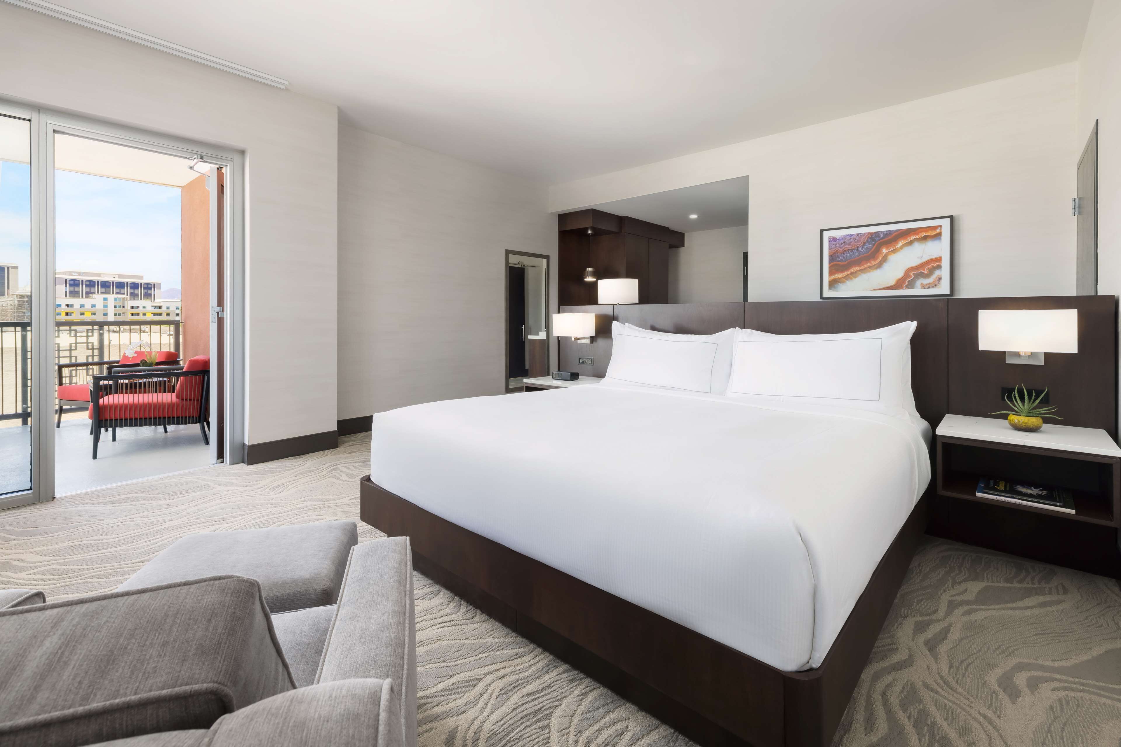 A hotel room with a large bed, a sitting area with a chair, a desk, and a small plant.