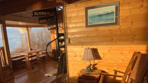 Een gezellig cabin-interieur met houten details, een wenteltrap, een eettafel, een stoel, een lamp en een lijst met een afbeelding aan de muur.