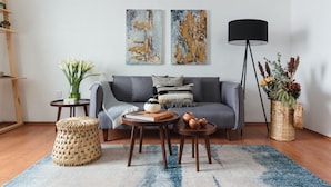 A modern living room with a grey sofa, two abstract paintings, a black floor lamp, a small round table with books and pottery, a woven pouf, and a blue rug.