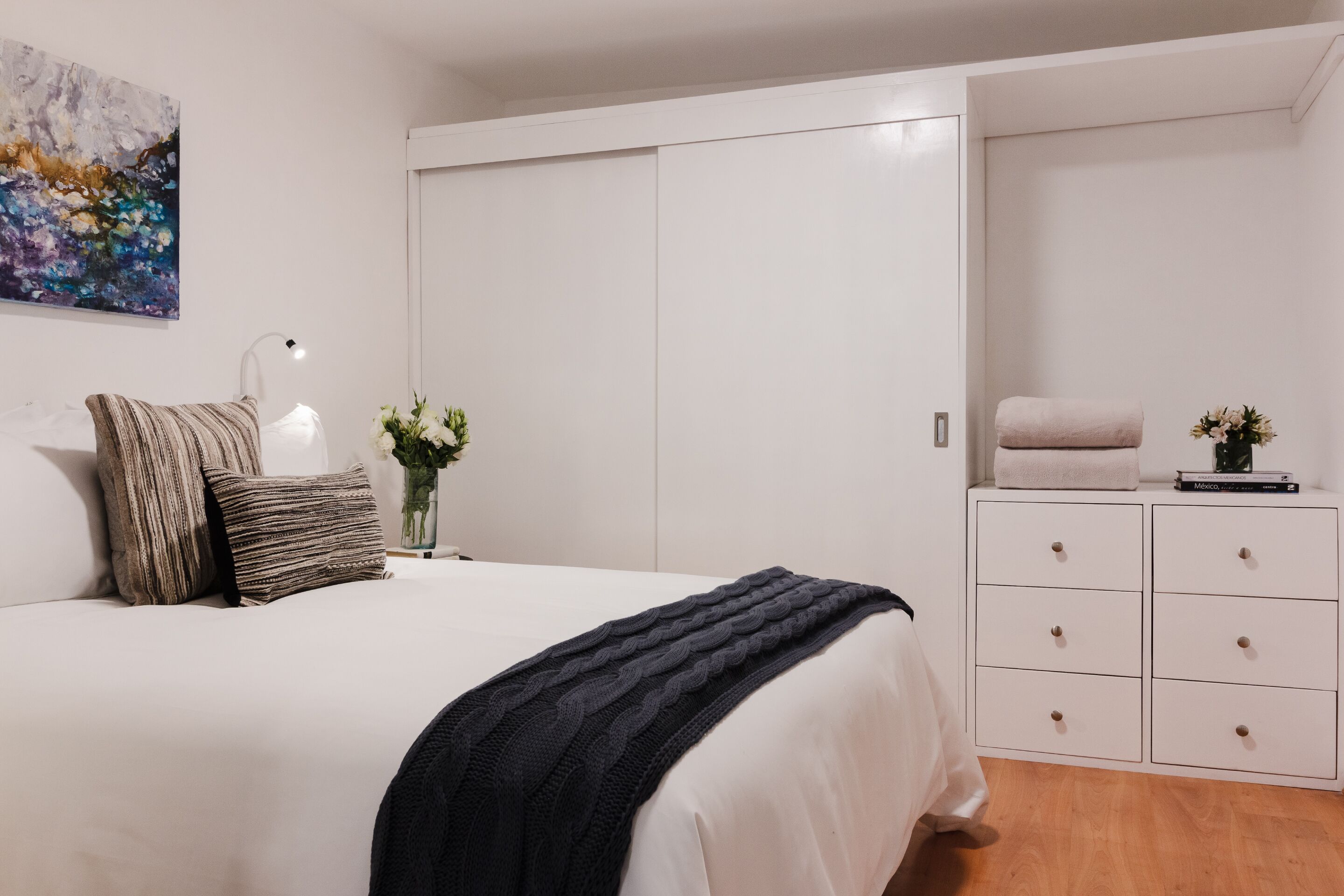 A bedroom with a large bed, a bedside table with a lamp, a dresser, and a painting on the wall.